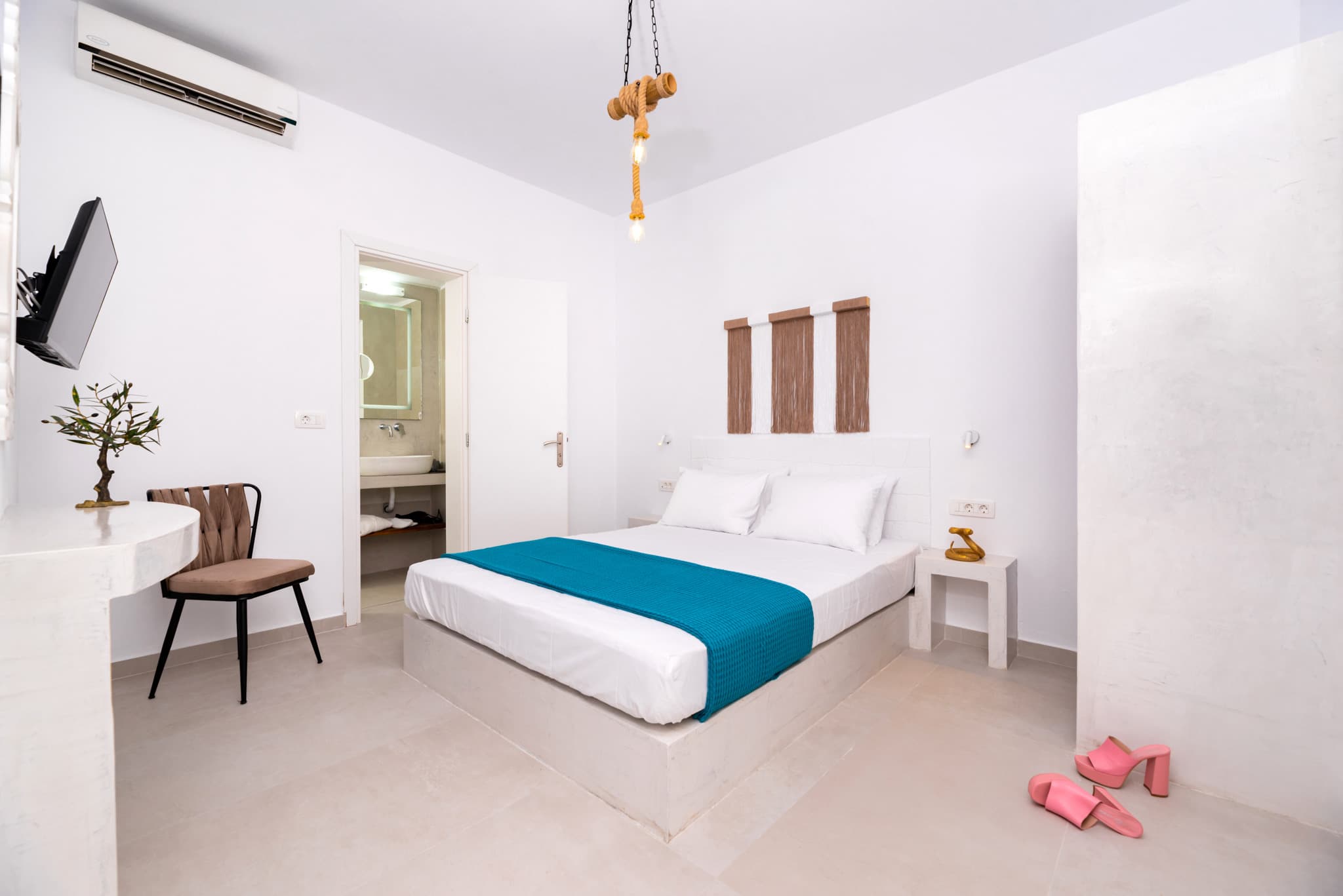 Minimalist white bedroom with blue bed runner, hanging rope light, and pink heels on floor.