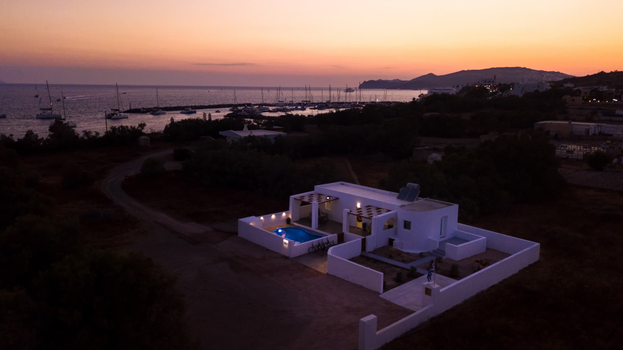 White villa with a glowing blue pool overlooks a harbor full of sailboats at sunset.